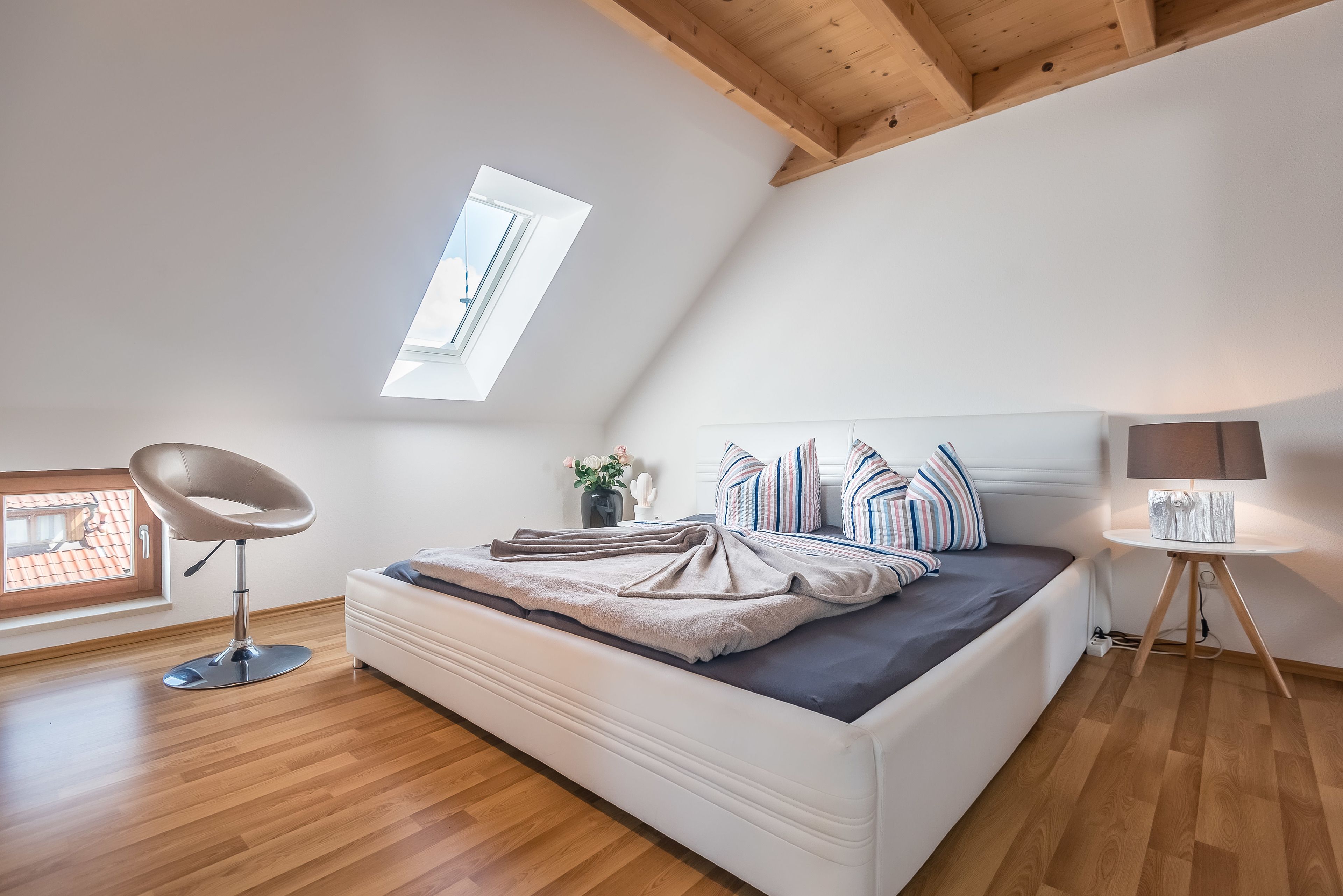 Une chambre avec un puits de lumière, un lit avec des coussins à rayures, une chaise moderne et une table de chevet avec une lampe.