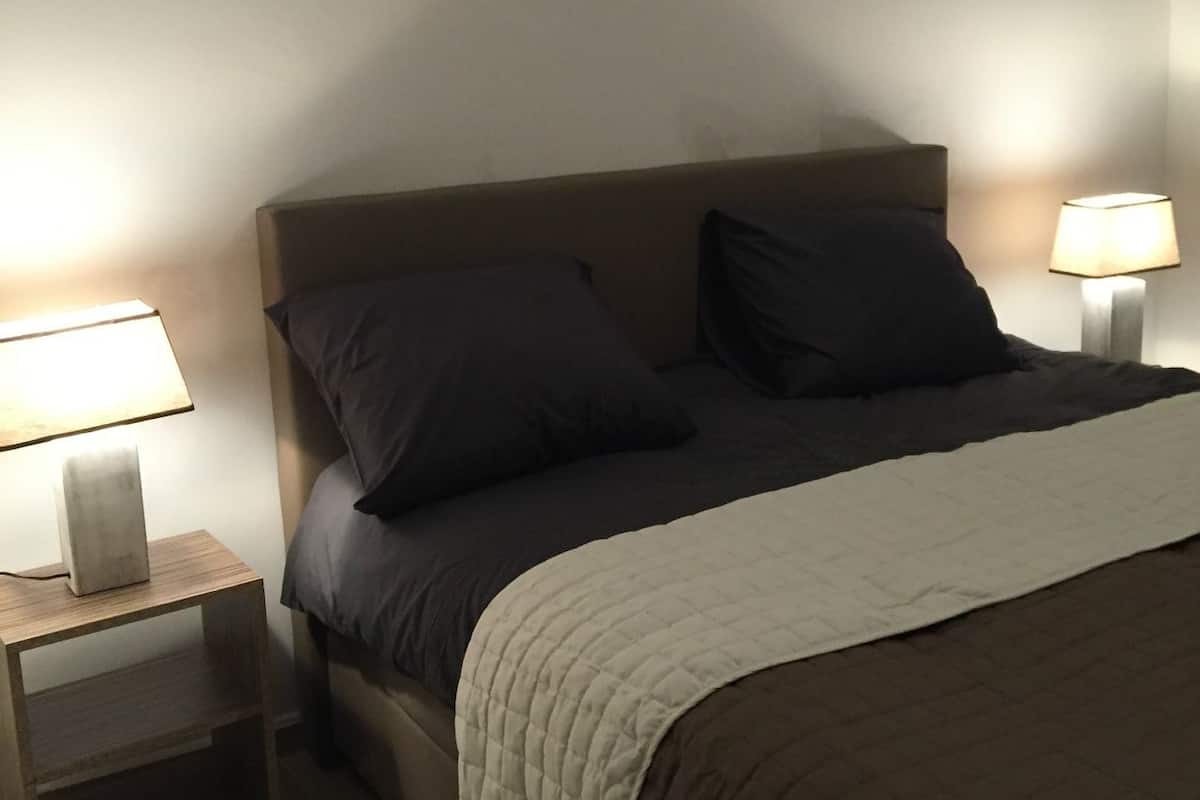 Una cama bien hecha con un cabecero oscuro, dos almohadas y una colcha acolchada. Hay dos mesitas de noche con lámparas, una a cada lado de la cama.