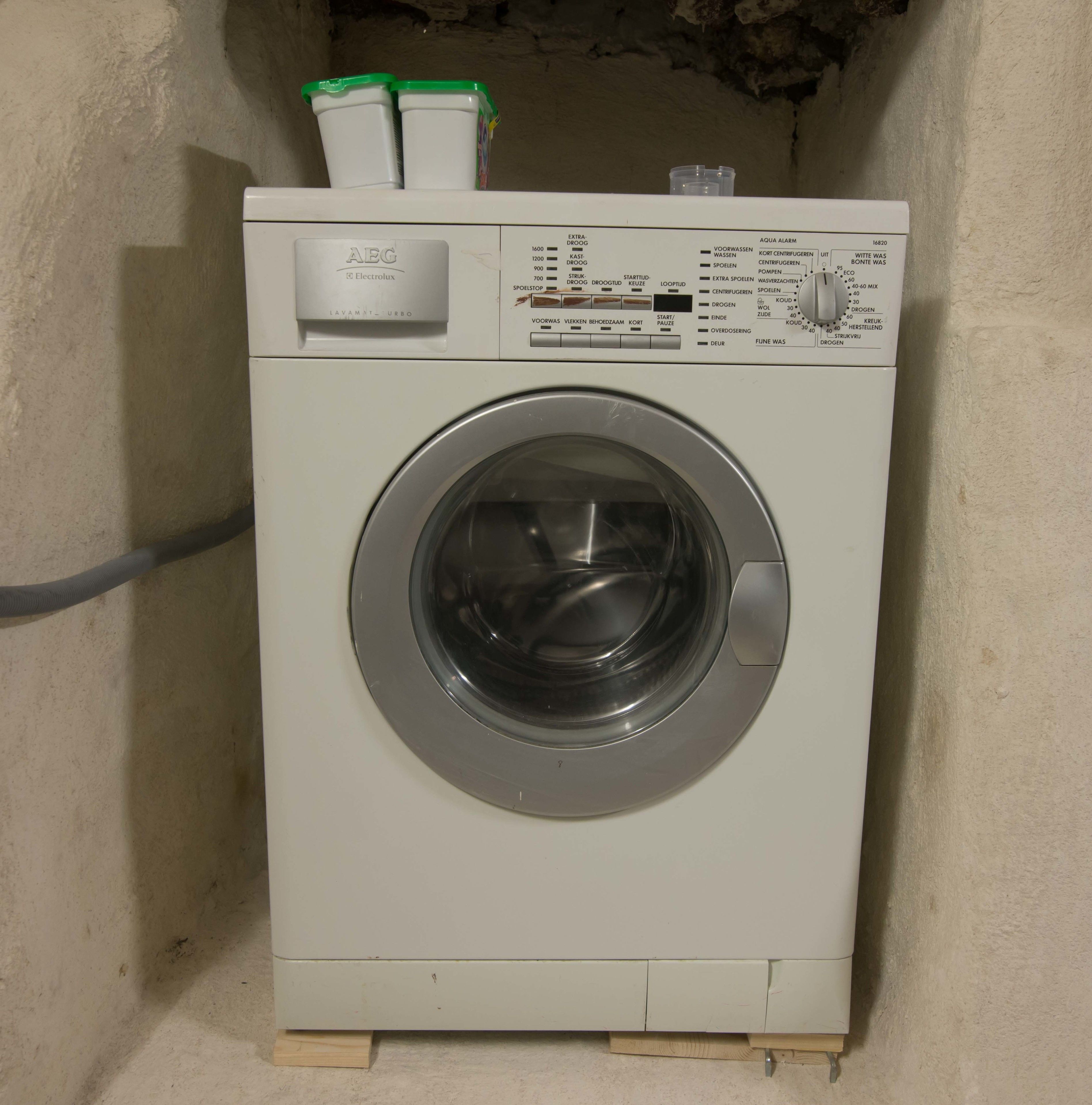 A front-loading washing machine with a control panel, placed in a corner with a wall-mounted shelf above it.