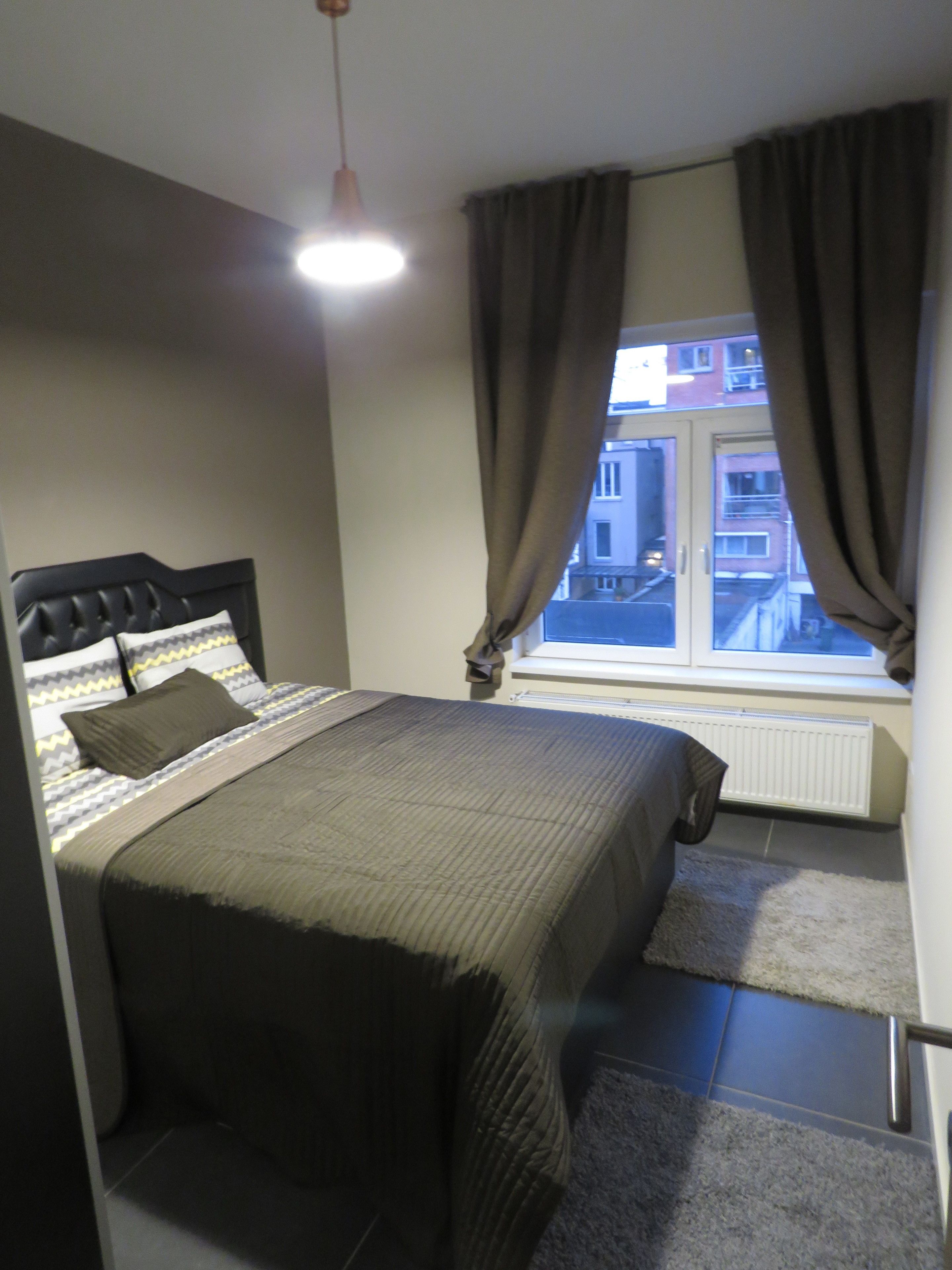Un dormitorio con cama, ventana con cortinas, radiador y vistas a edificios.