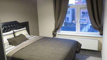 Un dormitorio con cama, ventana con cortinas, radiador y vistas a edificios.