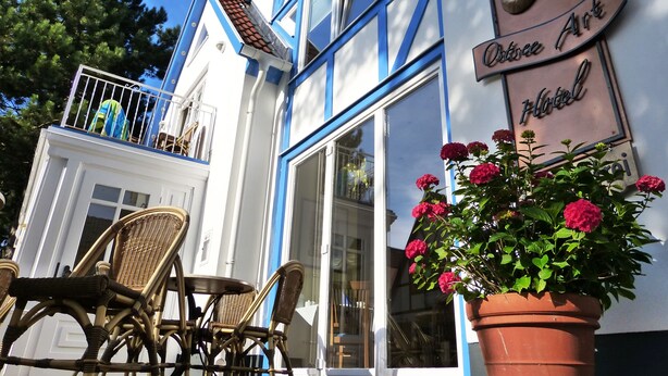 A hotel exterior with a balcony, wicker furniture, and a potted plant with flowers. - Vogel Hotel Appartements & Spa (Rostock)