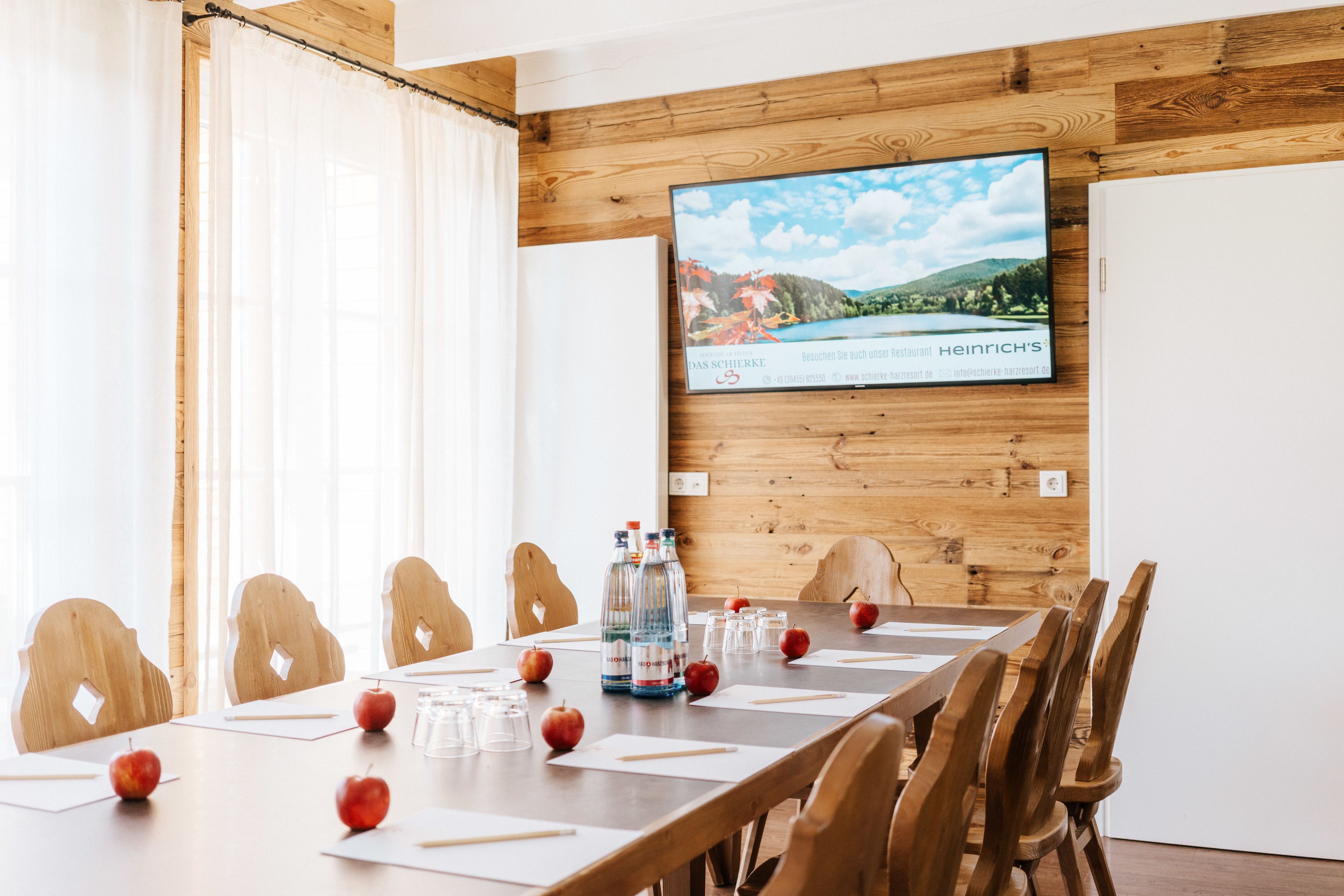 A conference room with a long table, wooden chairs, a TV screen, and a scenic view on the wall.