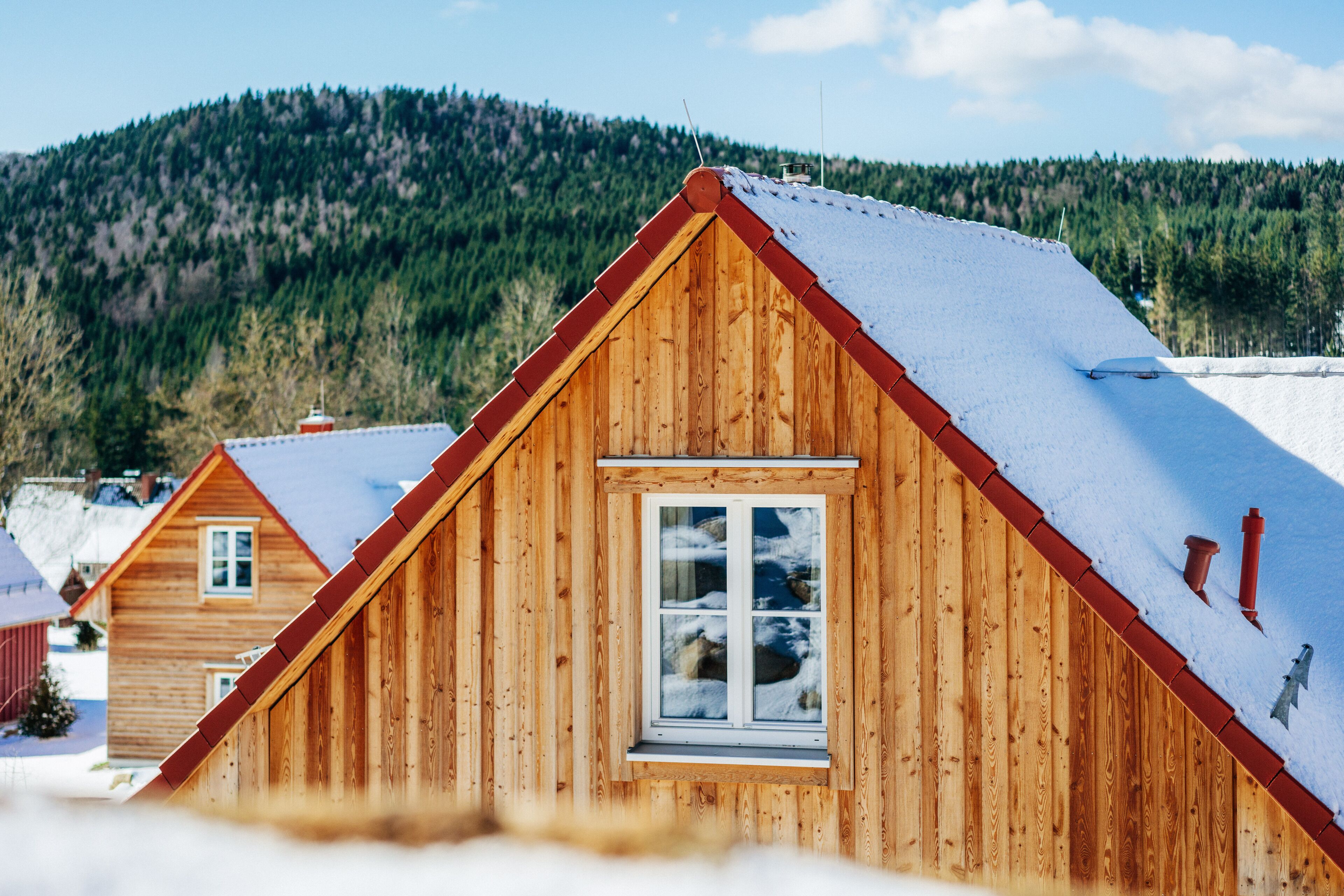 Una cabaña de madera con techo a dos aguas y una ventana, rodeada de nieve y una montaña boscosa al fondo.