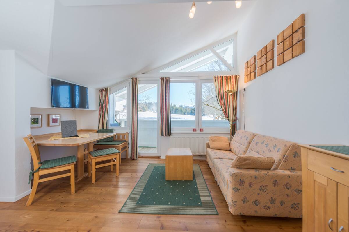Een gezellige woonkamer met een houten tafel, stoelen, een bank en een televisie die aan de muur is bevestigd.