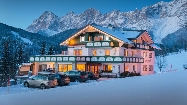 A multi-story hotel with a snow-covered mountain backdrop, featuring a parking area with several cars. - Landhaus Bergrast (Ramsau am Dachstein)