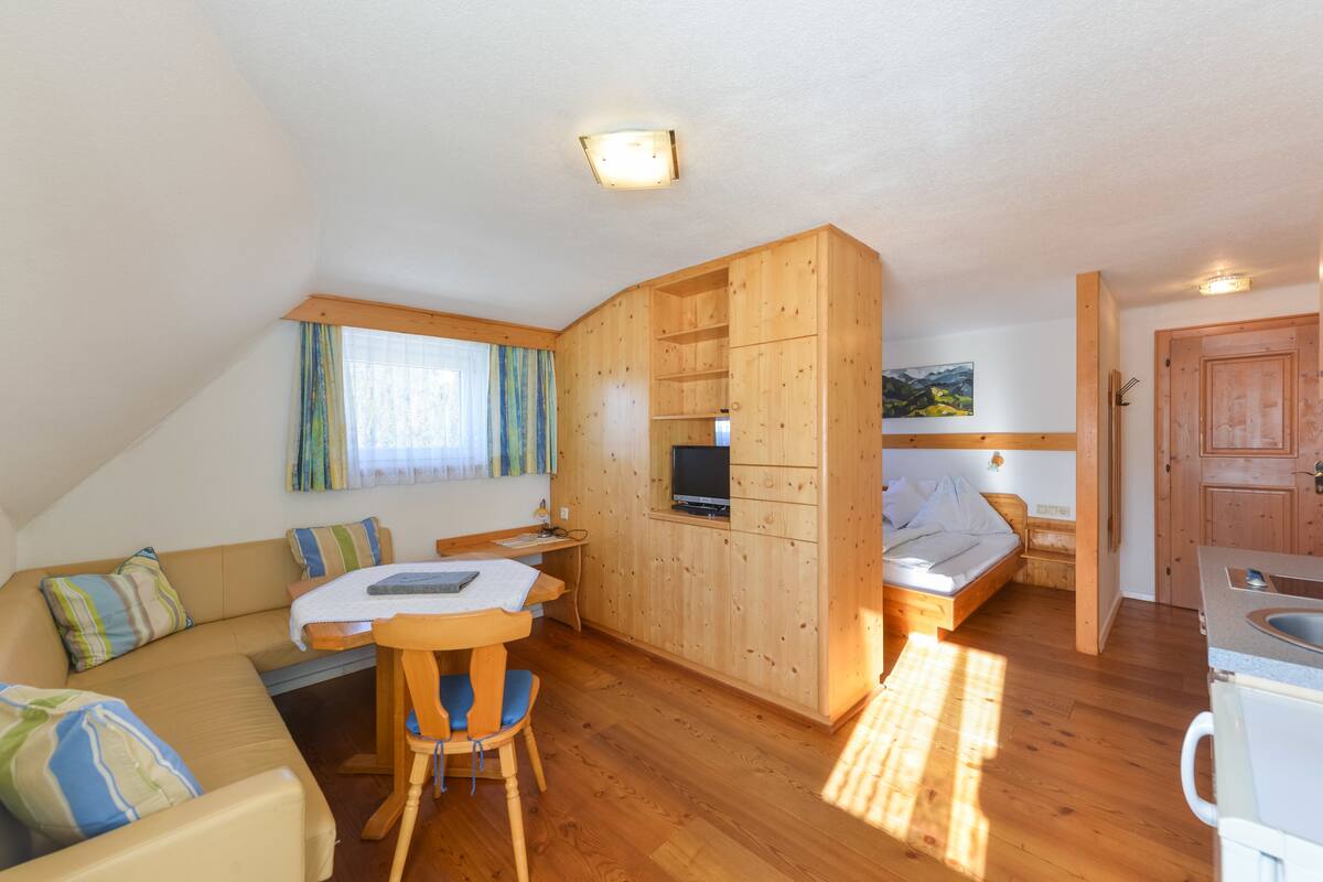 Een compacte kamer met een houten plafond, een bed, een televisie, een eettafel met stoelen en een kitchenette.