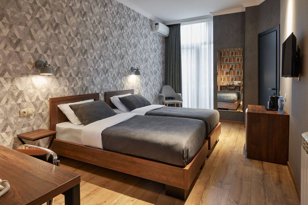 A modern hotel room with two beds, a wooden desk, a bookshelf, and a TV.