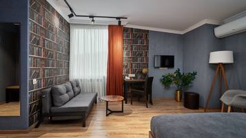 A modern living room with a grey sofa, a wooden coffee table, a bookshelf, a desk with a chair, a potted plant, and a lamp.