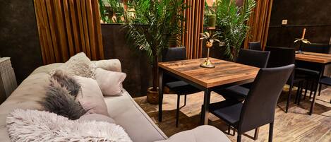 A modern dining area with a wooden table and black chairs, surrounded by a sofa with fluffy cushions and a decorative plant.