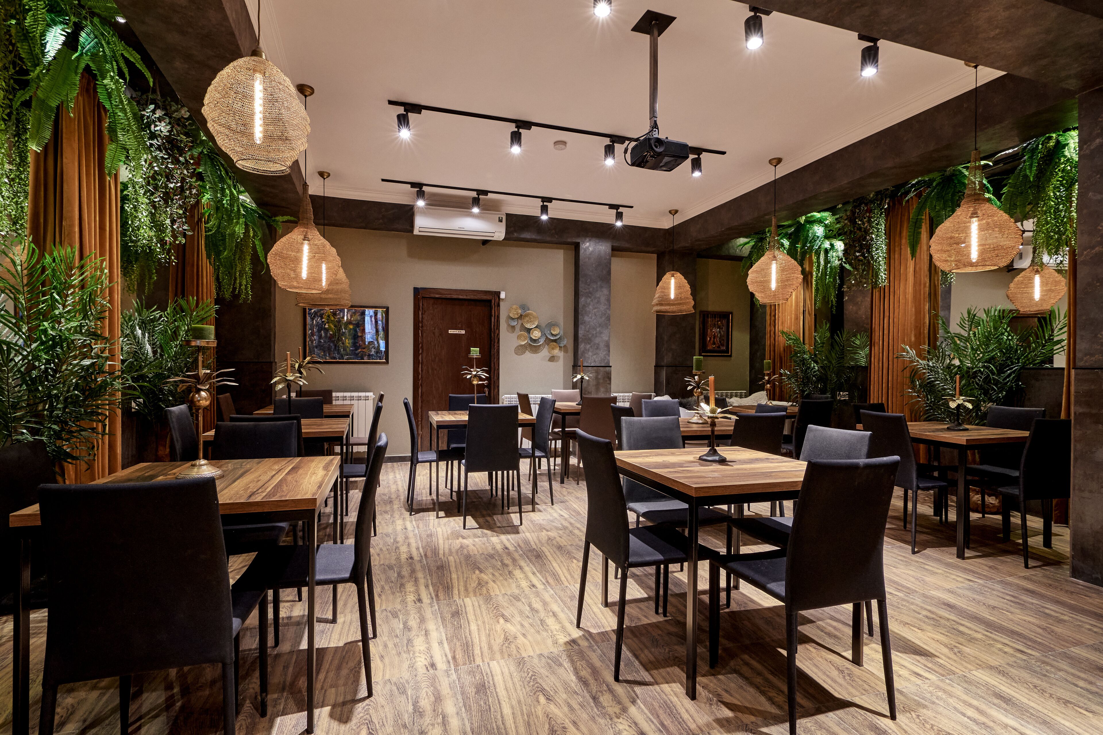 A restaurant with wooden tables and chairs, hanging pendant lights, and lush green plants.