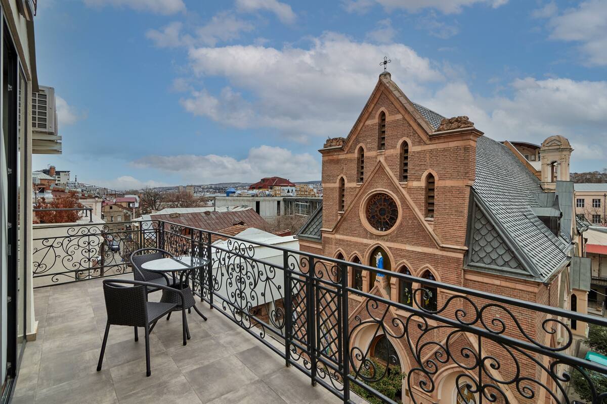 A balcony with a wrought iron railing, a table, and two chairs. In the background, there is a brick building with a pointed roof and a circular window.