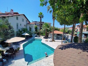 A swimming pool with lounge chairs and umbrellas, surrounded by buildings and palm trees. -  Göcek Selya Hotel (Fethiye)