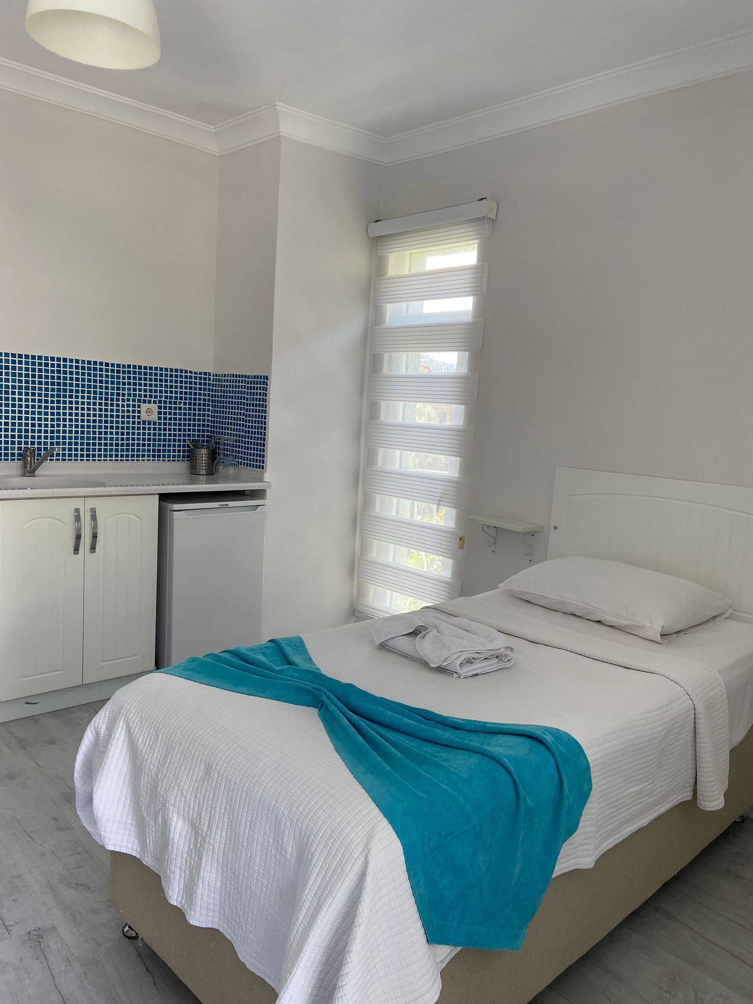 A bedroom with a bed, a nightstand, a window with blinds, and a kitchenette with a blue tiled backsplash.