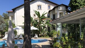 Zona piscina con lettini e ombrelloni, circondata da piante tropicali e una casa grande sullo sfondo.