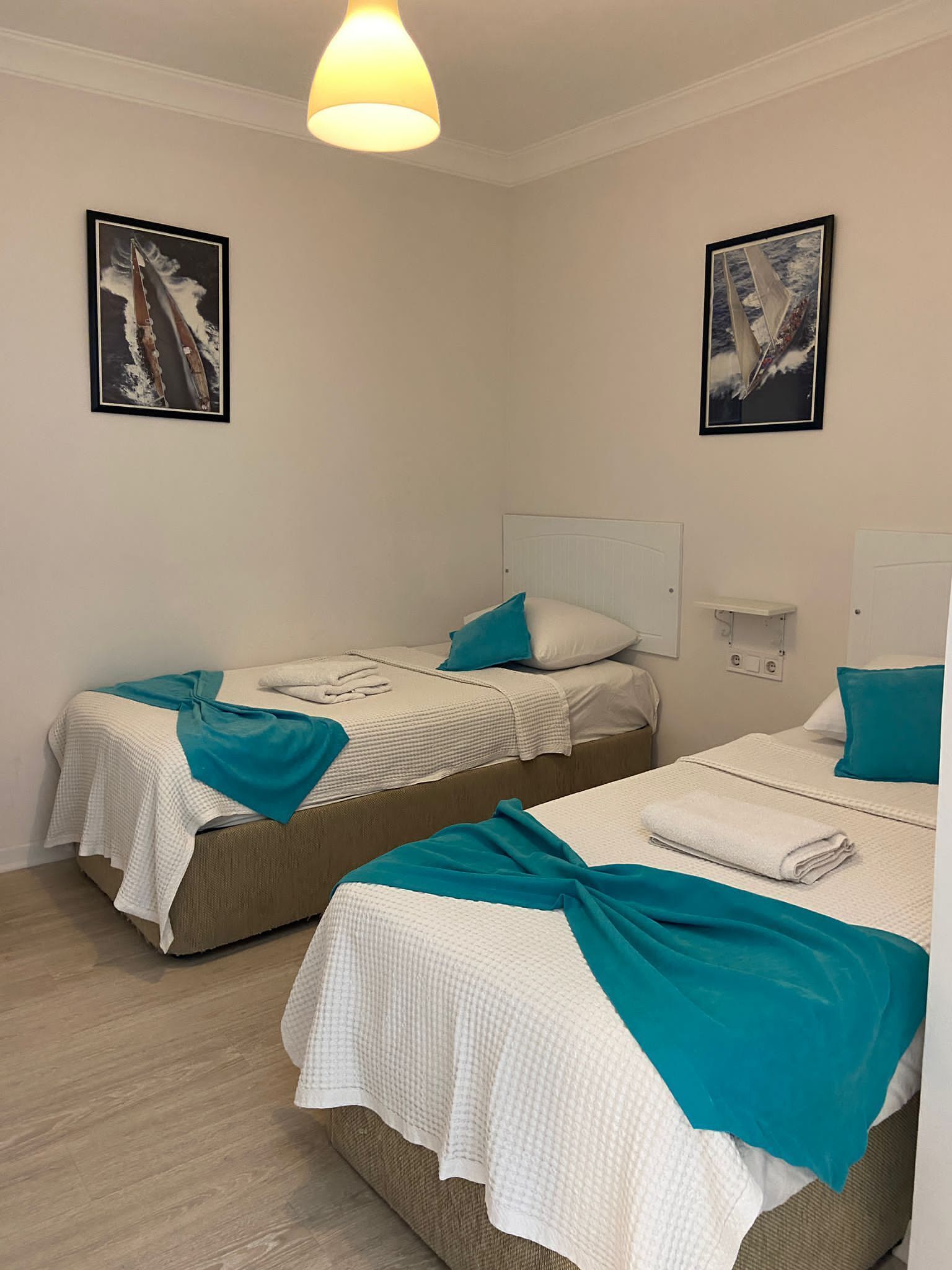A hotel room with two beds, each with a teal blanket and pillow, white bedding, and a headboard. There are two framed pictures on the walls, a ceiling light, and a bedside table with a lamp.