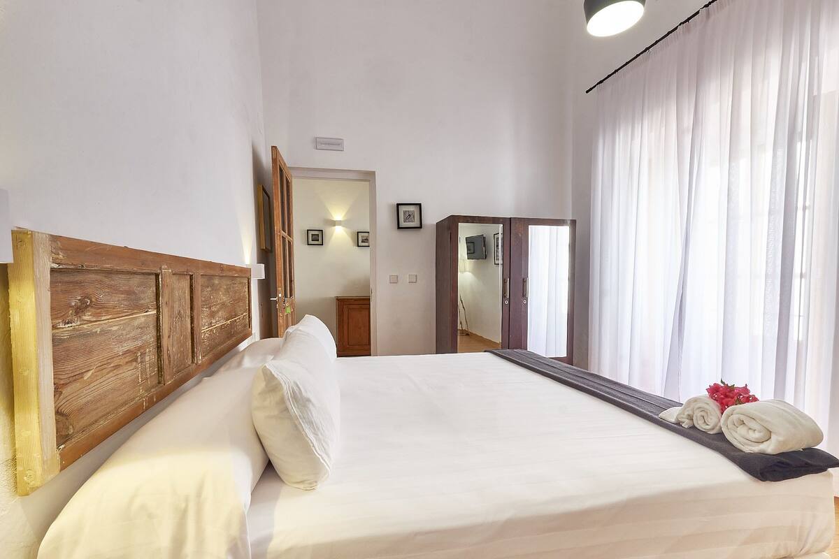 A hotel room with a wooden headboard, a bed with white linens, a nightstand, a mirror, a wardrobe, and a window with sheer curtains.