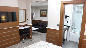 Habitación de hotel con cama, escritorio con silla, sofá, zona de ducha y televisión montada en la pared.