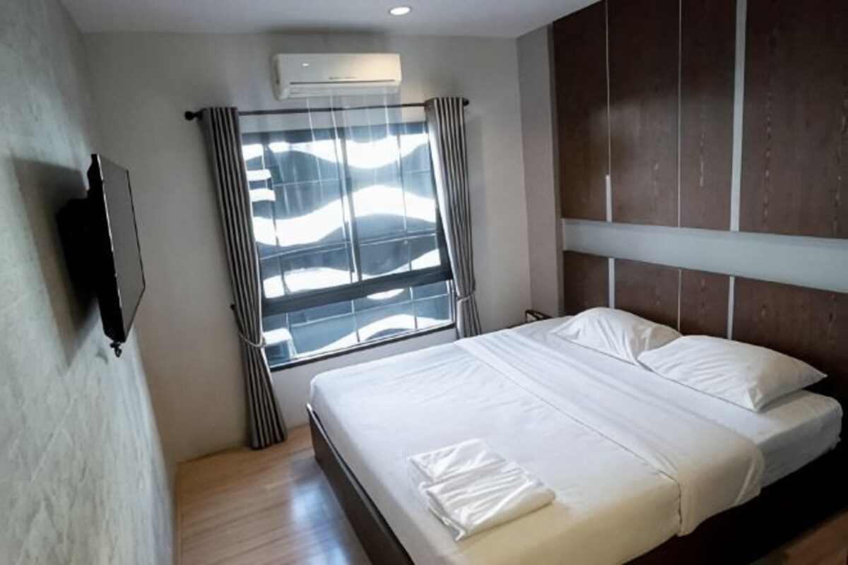A hotel room with a bed, a TV mounted on the wall, a window with curtains, and a wooden floor.