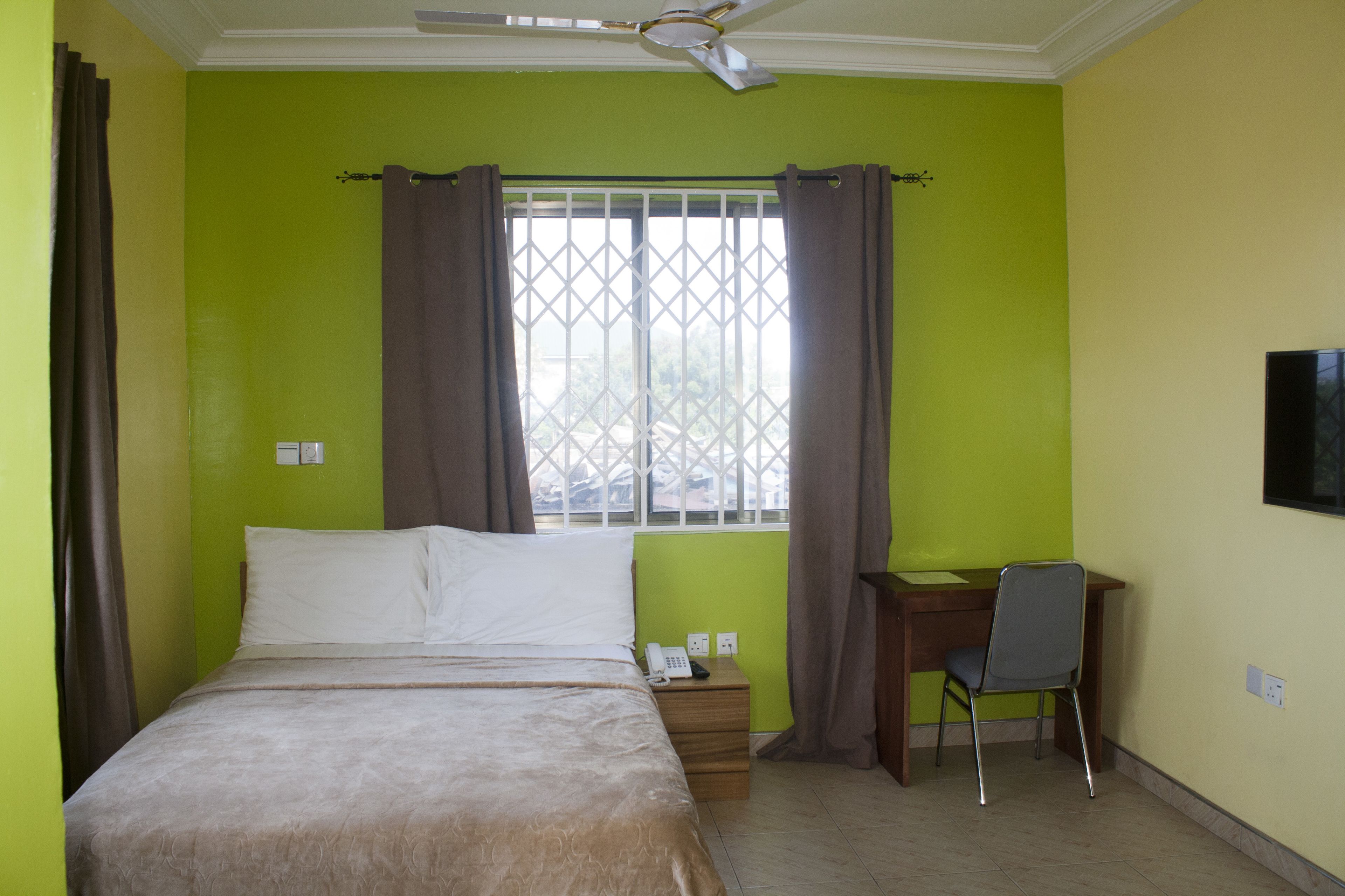 A bedroom with a bed, a desk, a chair, a window with curtains, and a ceiling fan.