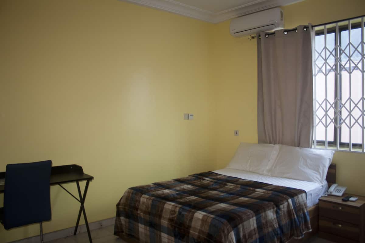 Quarto com cama, escrivaninha, cadeira e janela com cortina.
