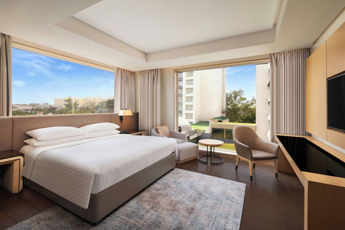 A modern hotel room with a large bed, a desk, a chair, and a TV. The room has large windows with curtains and a view of buildings outside.