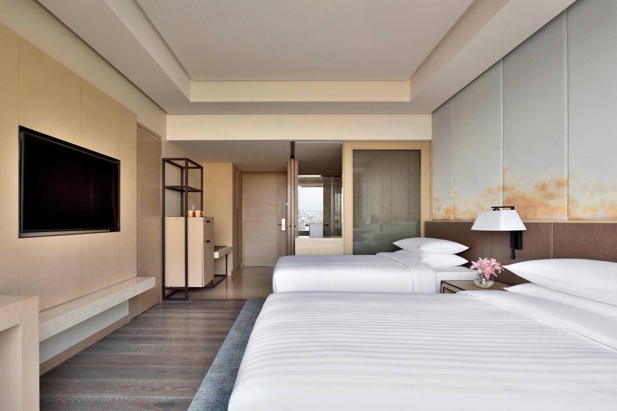A hotel room with two beds, a TV, a bathroom with a mirror, and a view of the city.