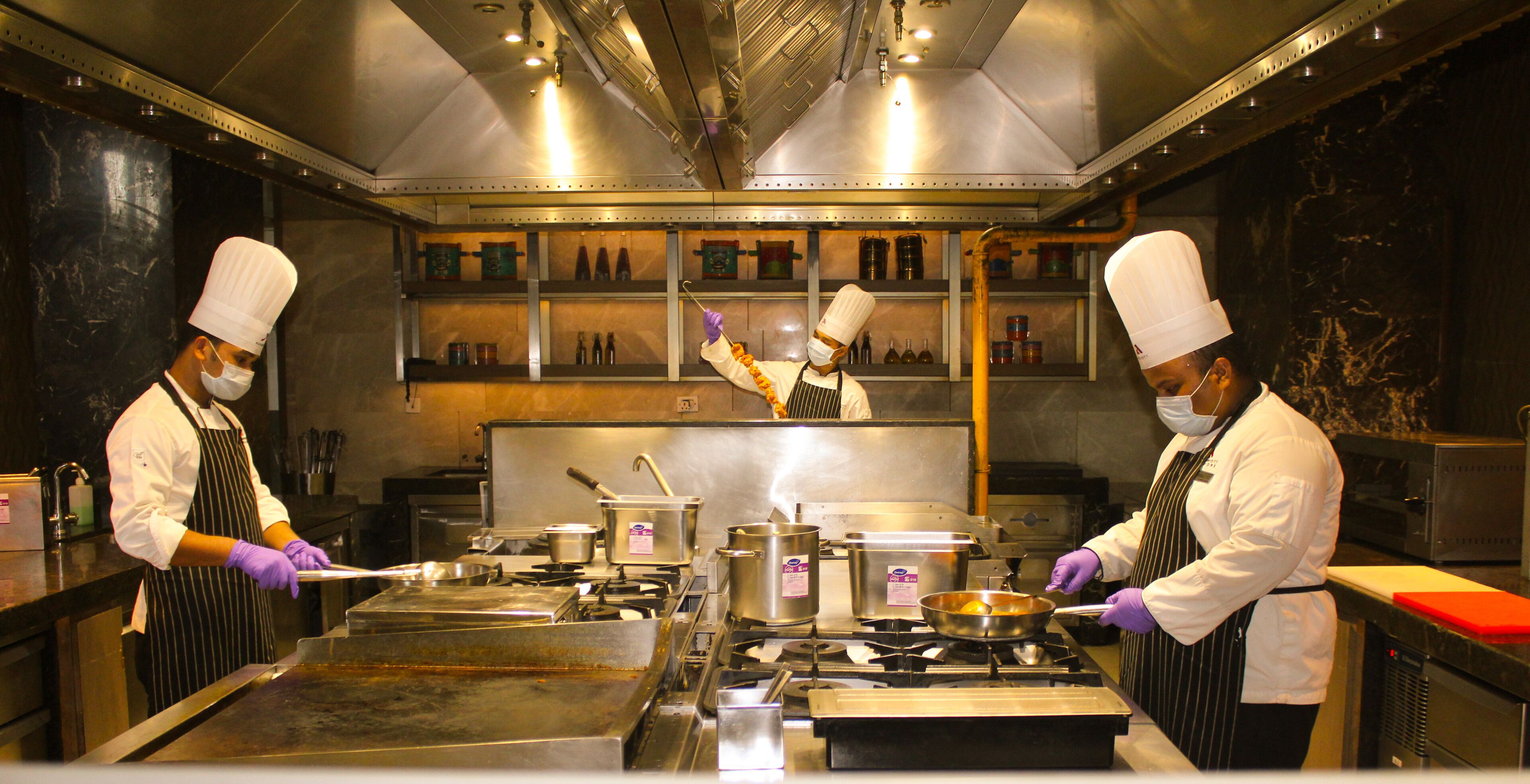 Una cocina profesional con electrodomésticos de acero inoxidable, chefs con uniforme y gorro blanco, y diversos utensilios y alimentos para cocinar.