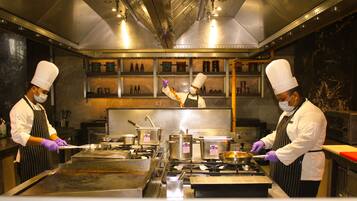 Una cocina profesional con electrodomésticos de acero inoxidable, chefs con uniforme y gorro blanco, y diversos utensilios y alimentos para cocinar.