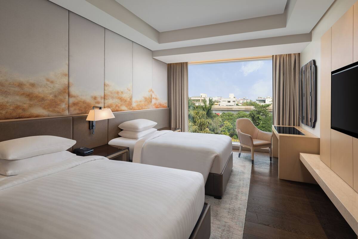A hotel room with two beds, a desk, a TV, and a view of the city.