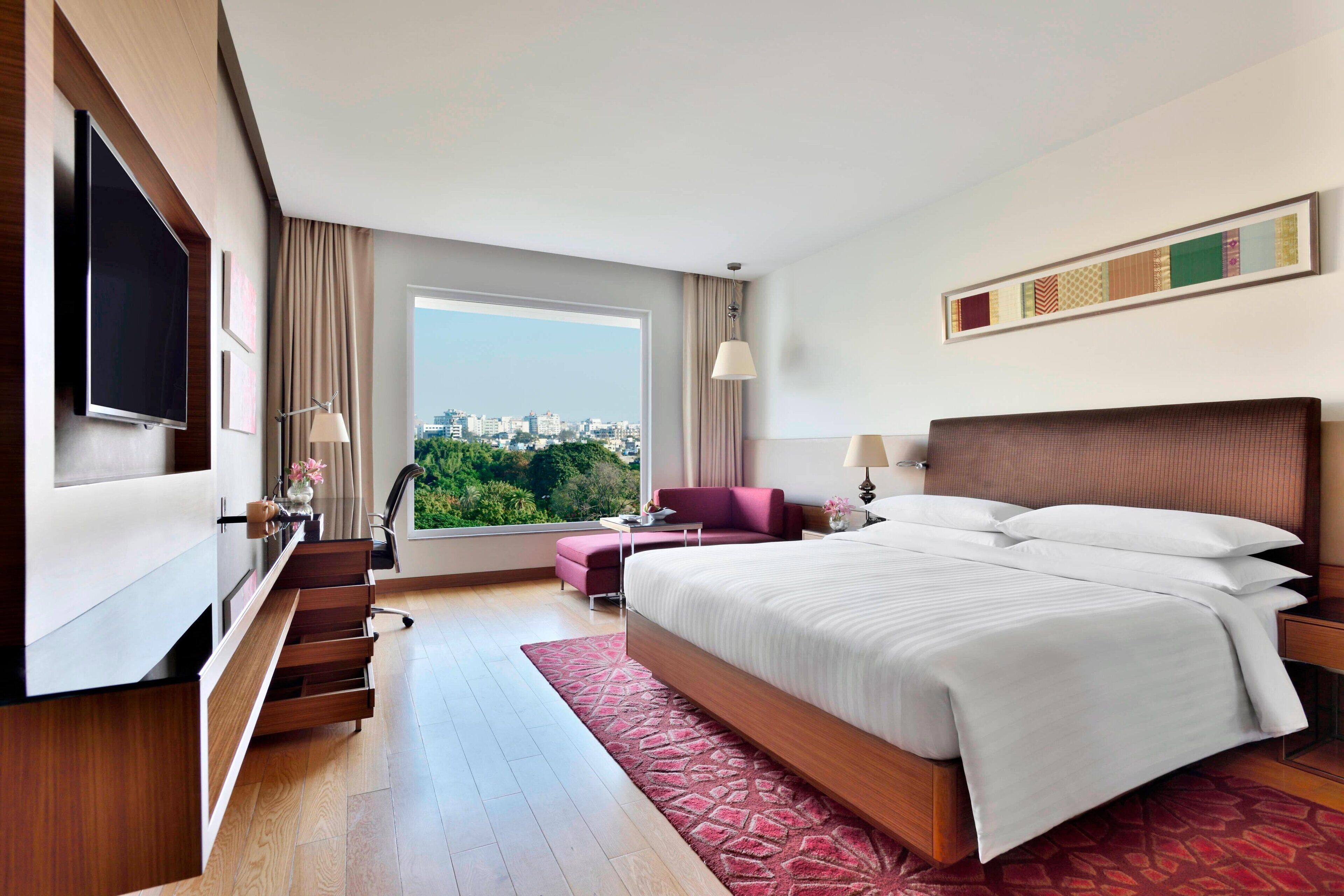 A modern hotel room with a large bed, a TV, a desk, and a view of the city.