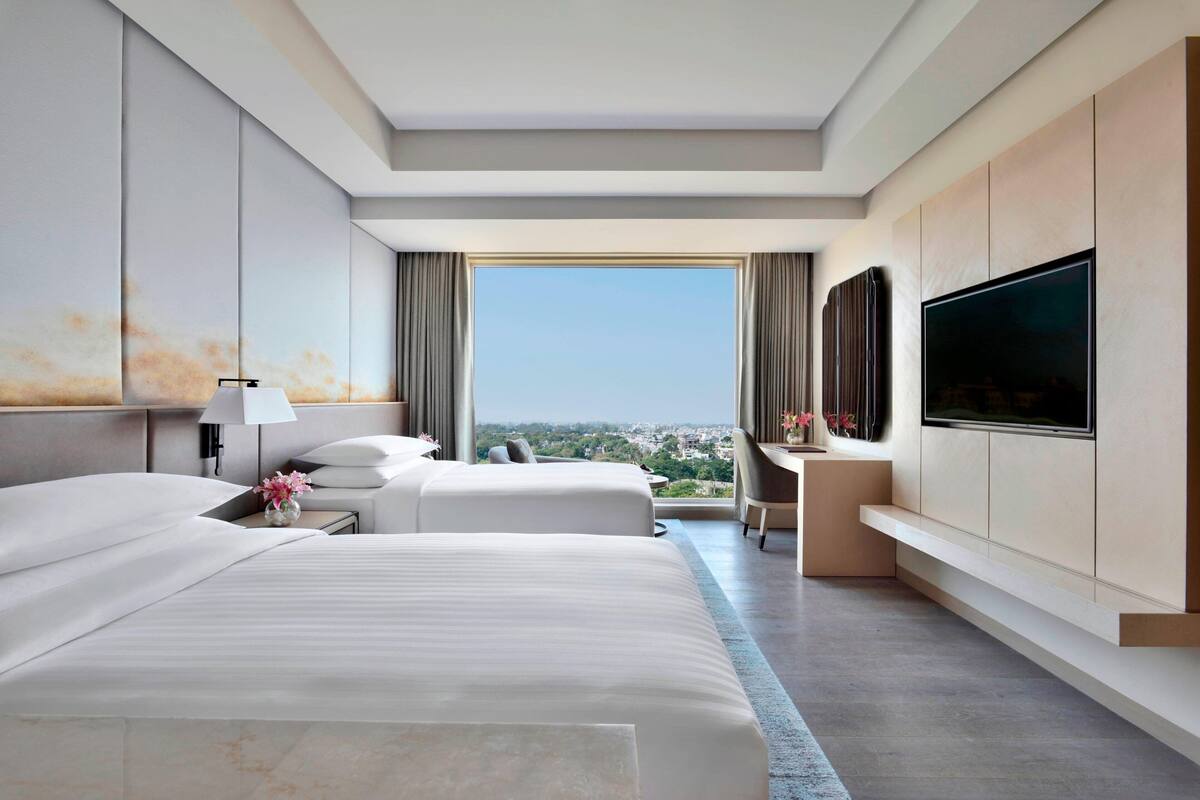 A modern hotel room with two beds, a large TV, and a view of the city.