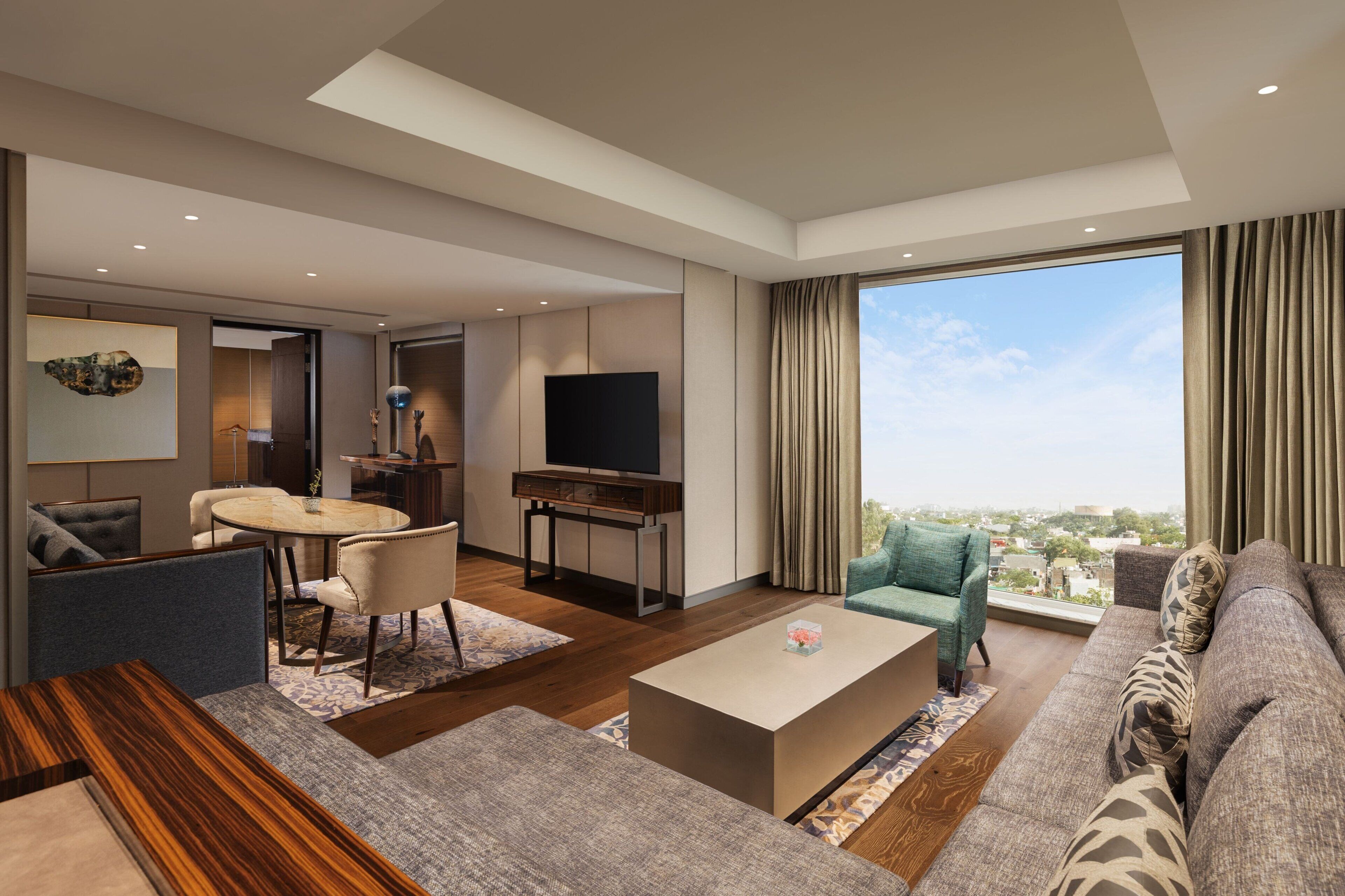 A modern living room with a sofa, coffee table, and TV. There is a dining area with a table and chairs. The room has large windows with curtains.