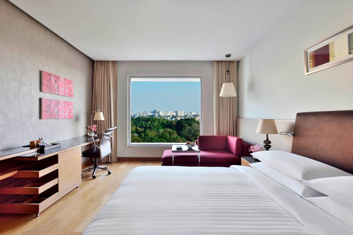 A hotel room with a large bed, a desk with a chair, a small table, a red sofa, and a view of a cityscape through a large window.