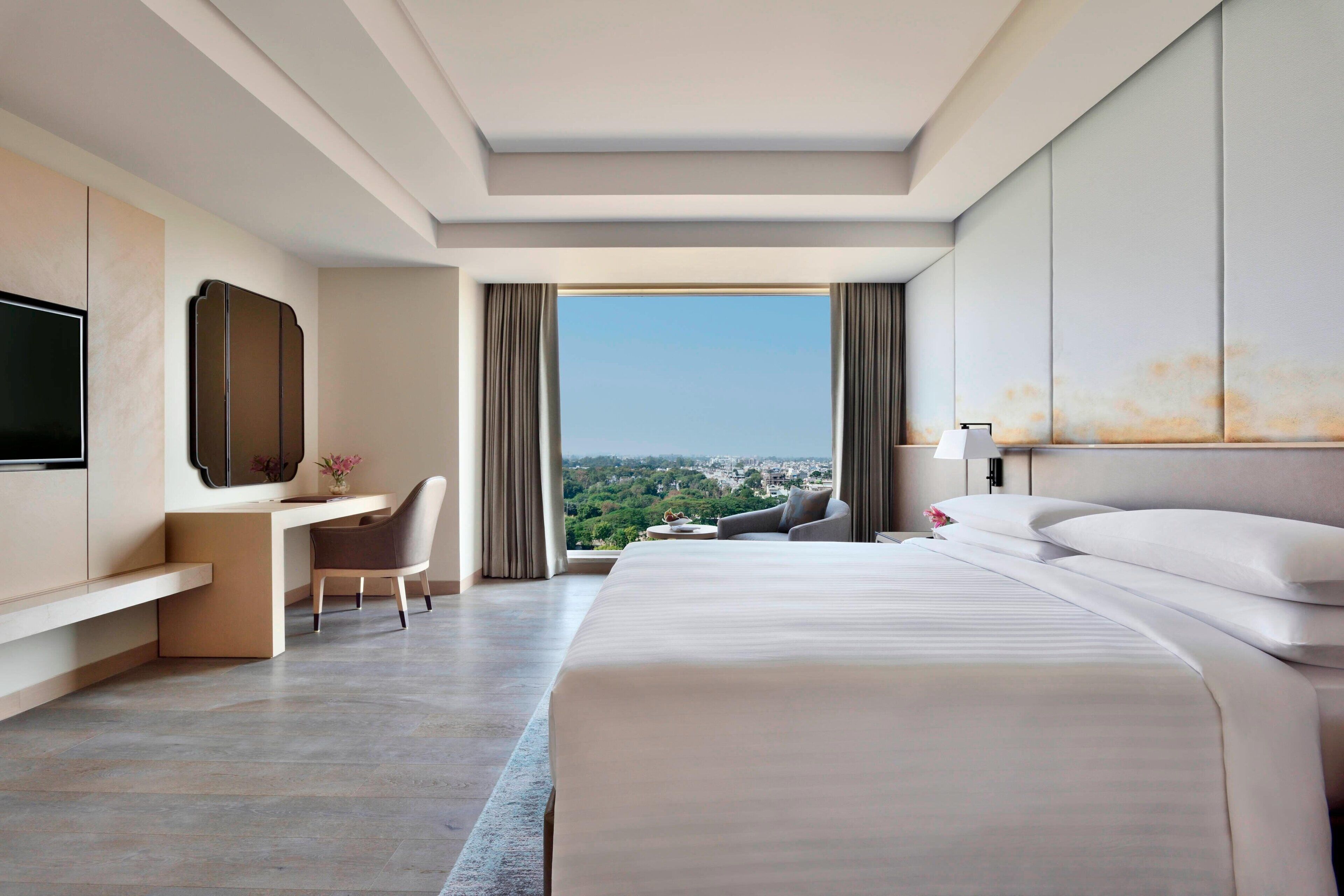 A modern hotel room with a large bed, a desk, a chair, a TV, and a view of the city.