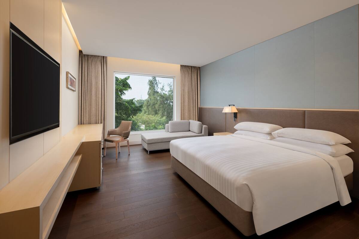 A modern hotel room with a large bed, a flat-screen TV, a seating area with a sofa and armchair, and a window with a view of greenery.
