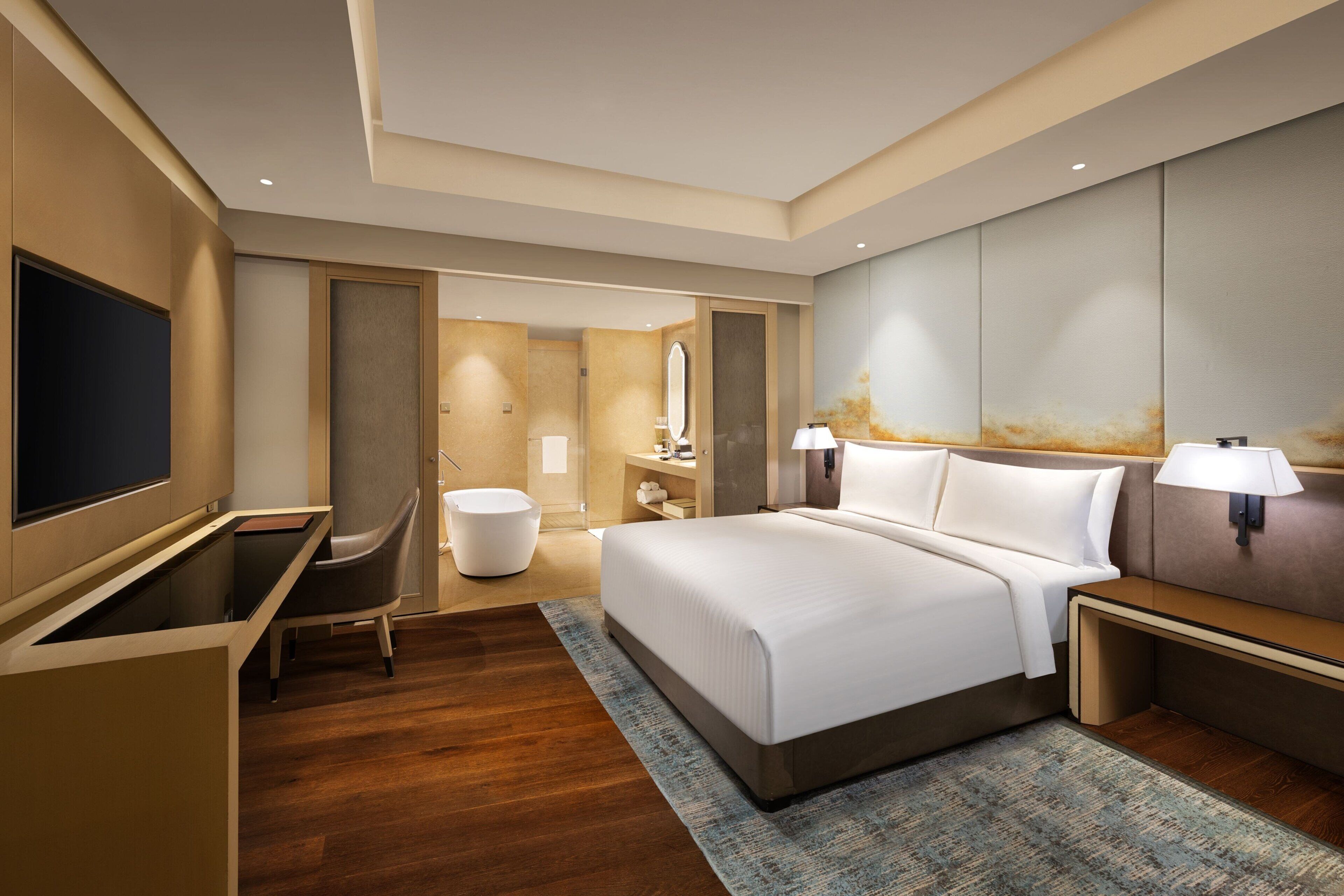 Une chambre d’hôtel moderne, dotée d’un grand lit, d’un bureau, d’une chaise, d’une télévision et d’une salle de bain visible à travers une porte ouverte.
