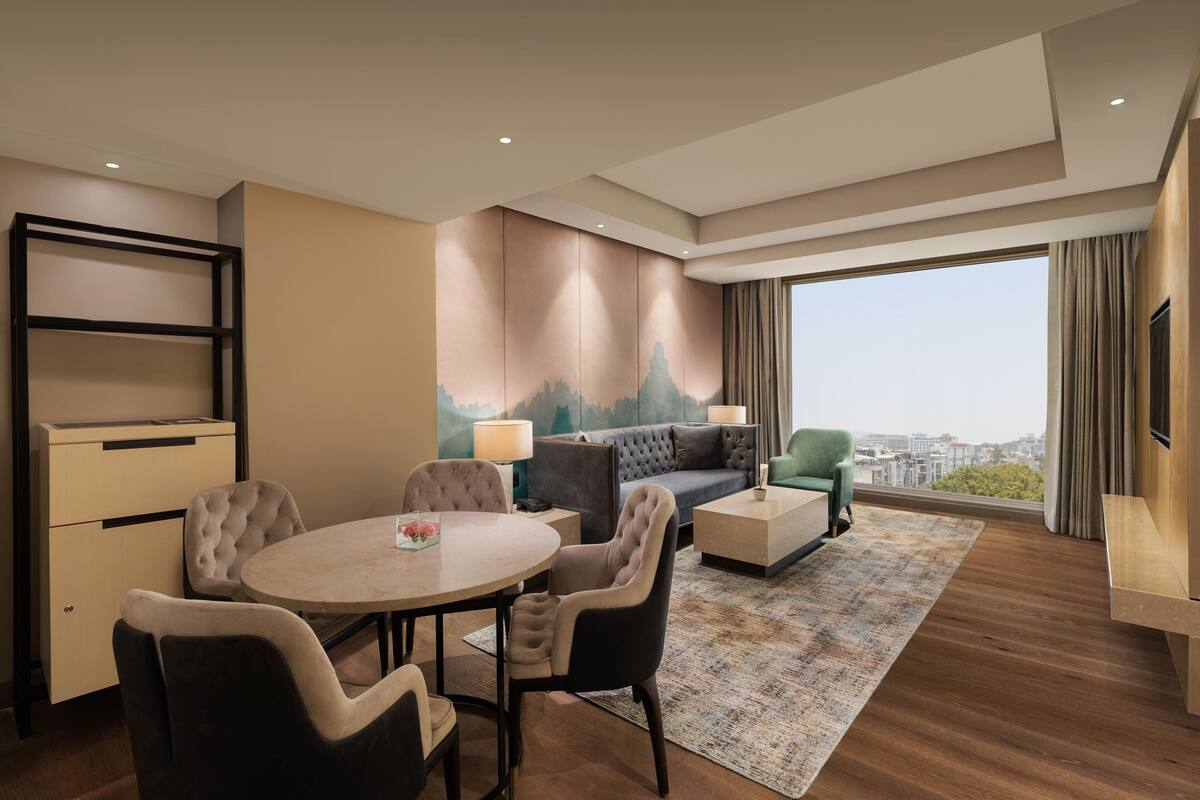 A modern hotel room with a dining area, a sofa, and a large window offering a city view.