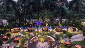 A well-lit outdoor dining area with multiple round tables and chairs, surrounded by lush greenery and palm trees.