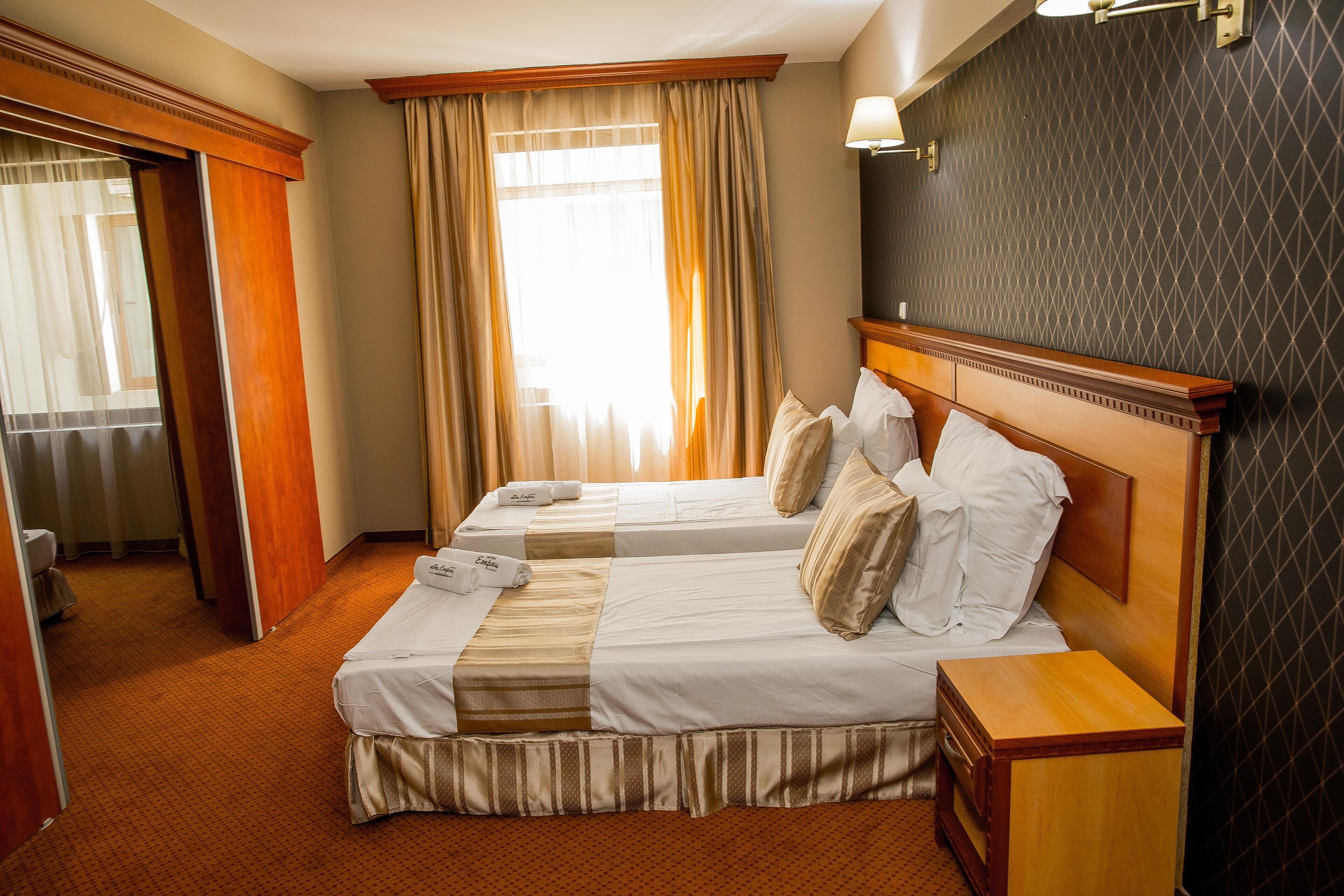 Quarto de hotel com duas camas, cabeceira de madeira, mesa de cabeceira e uma janela grande com cortinas.