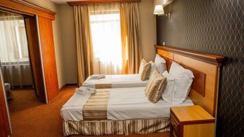 Quarto de hotel com duas camas, cabeceira de madeira, mesa de cabeceira e uma janela grande com cortinas.
