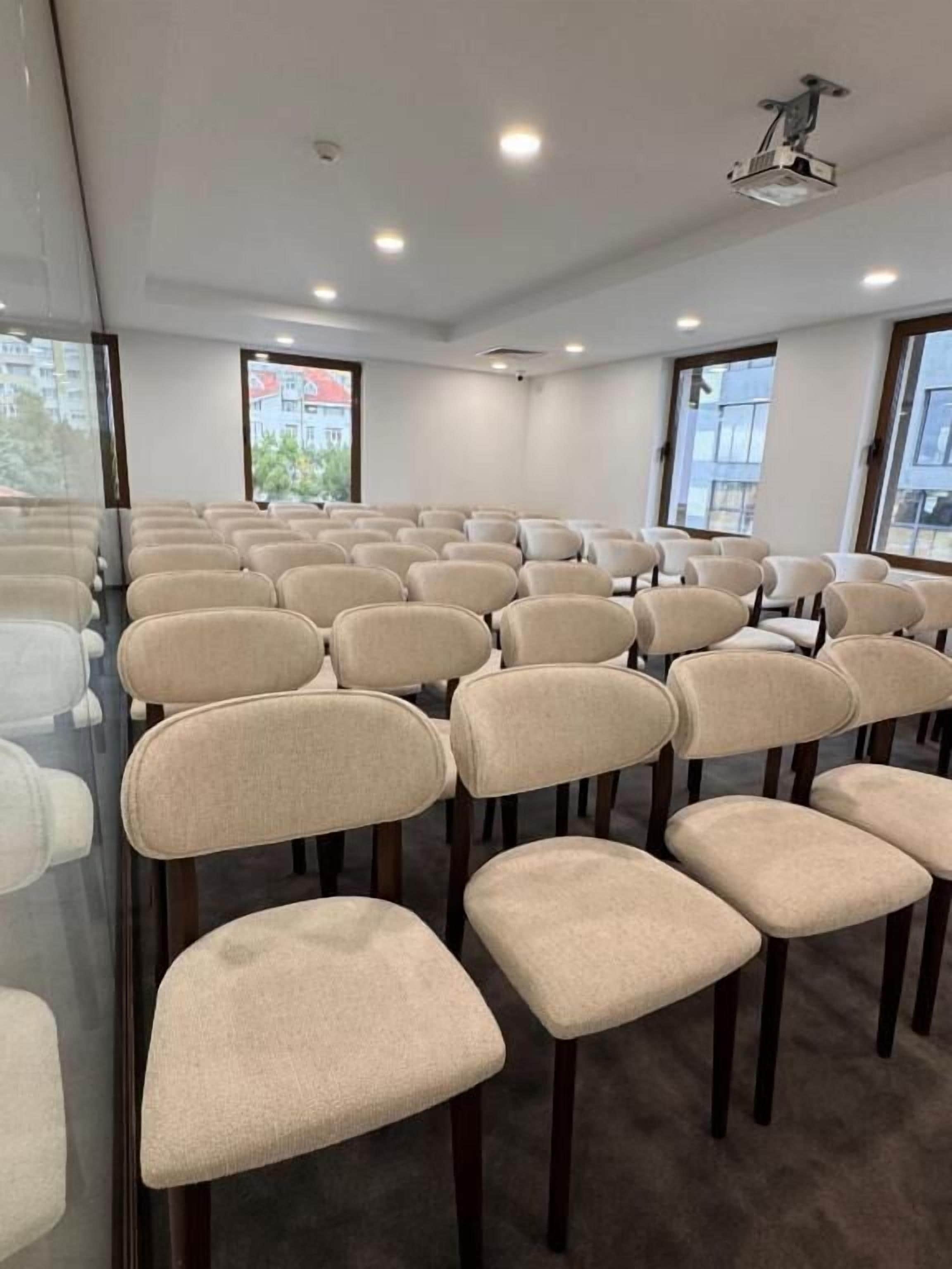 A conference room with rows of beige chairs, large windows, and ceiling-mounted projectors.