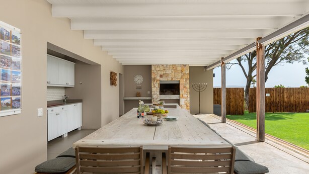 A spacious patio with a wooden table and chairs, a stone fireplace, and a view of a green lawn. - Sunset Splendour Guesthouse (Cape Town)