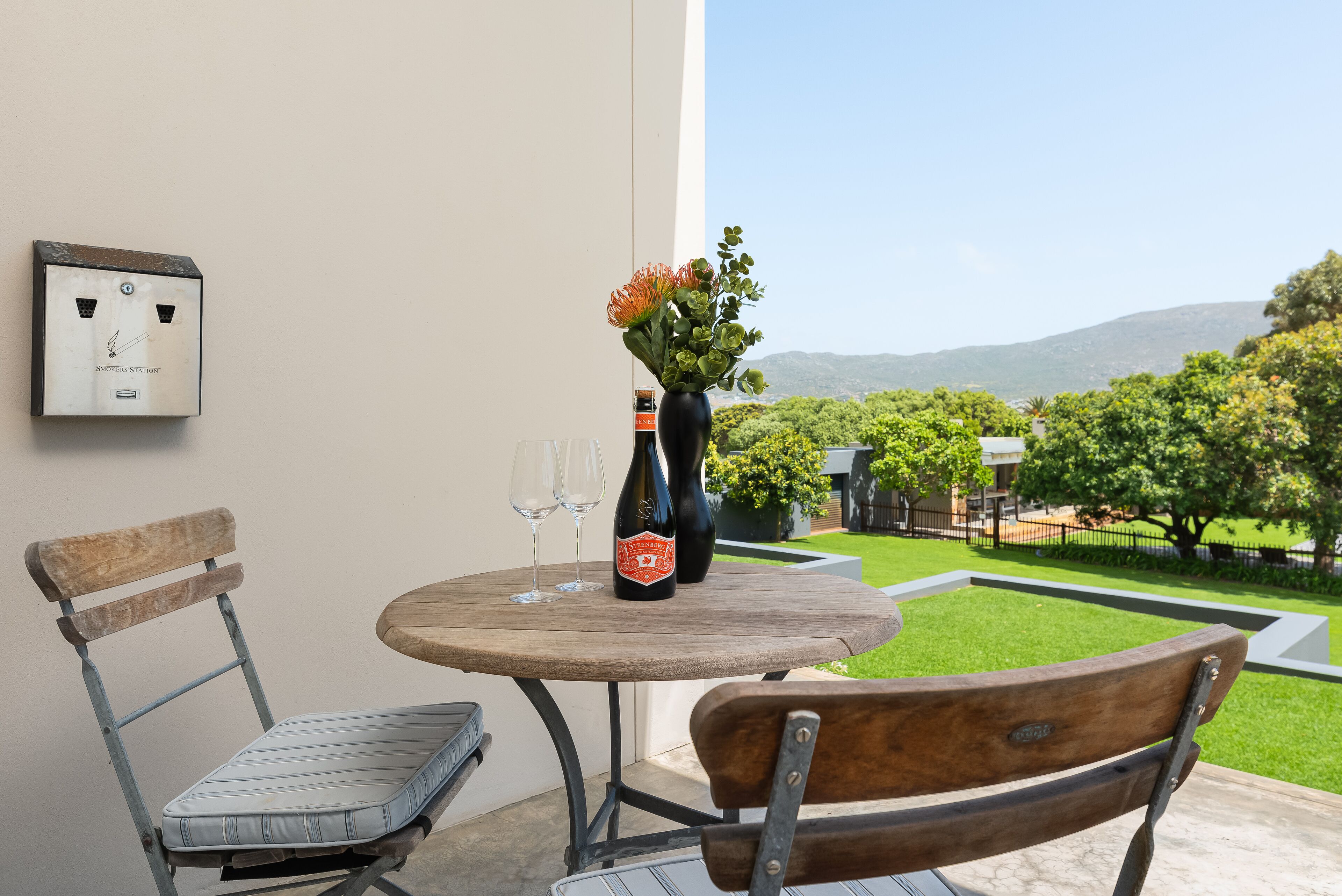 Een terras met een tafel, stoelen, een wijnfles en een vaas met bloemen.
