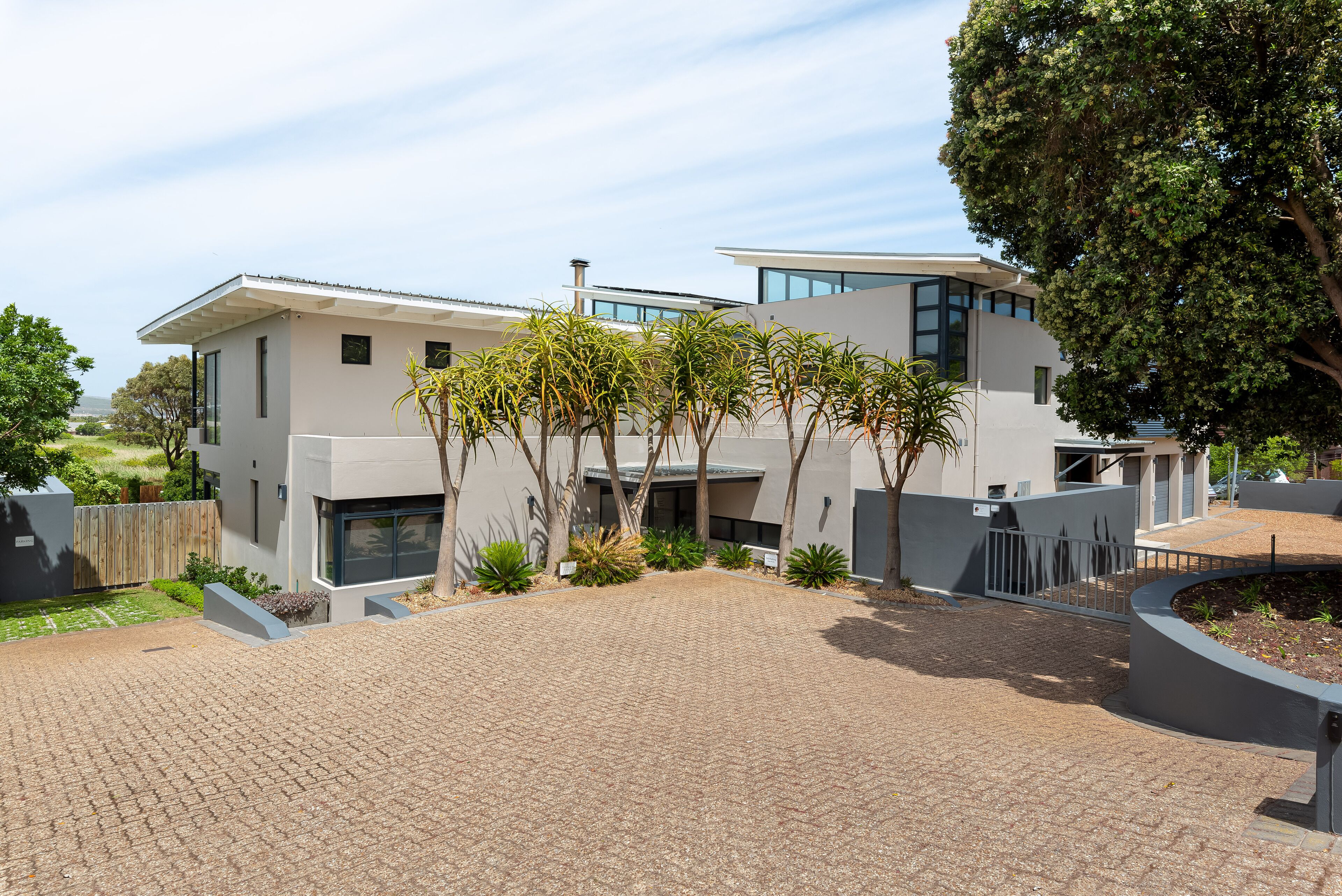 A modern two-story house with a paved driveway, surrounded by trees and a fenced garden.