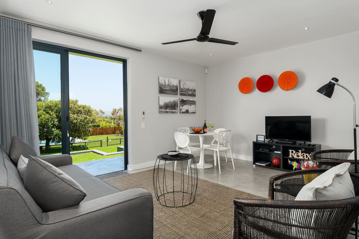 Uma sala de estar moderna com um sofá cinzento, uma mesa de centro redonda, uma área de jantar com mesa e cadeiras brancas, uma televisão de tela plana e uma varanda com vista para o verde.