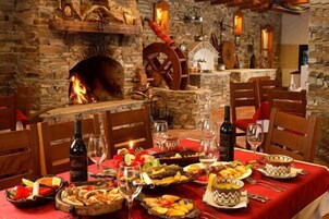 Un comedor con chimenea de piedra, sillas de madera y una mesa dispuesta con copas de vino, platos de comida y un mantel rojo.