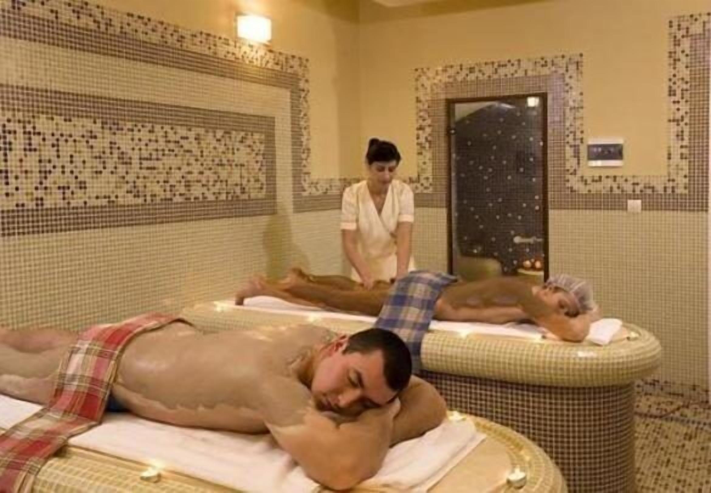 Ein Spa-Raum mit zwei Massagetischen, einer Person, die Klienten betreut, und einer gefliesten Wand.