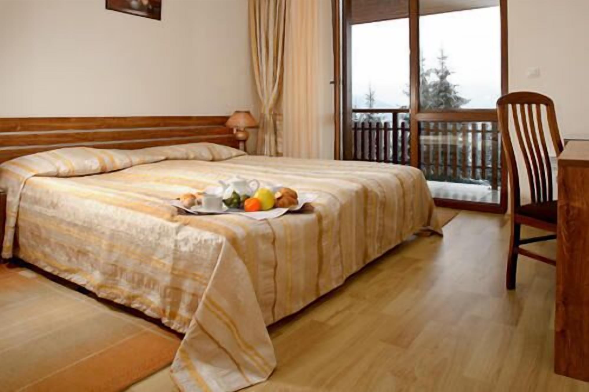 A hotel room with a bed, a dining table with fruits, a chair, and a balcony with a view of trees.