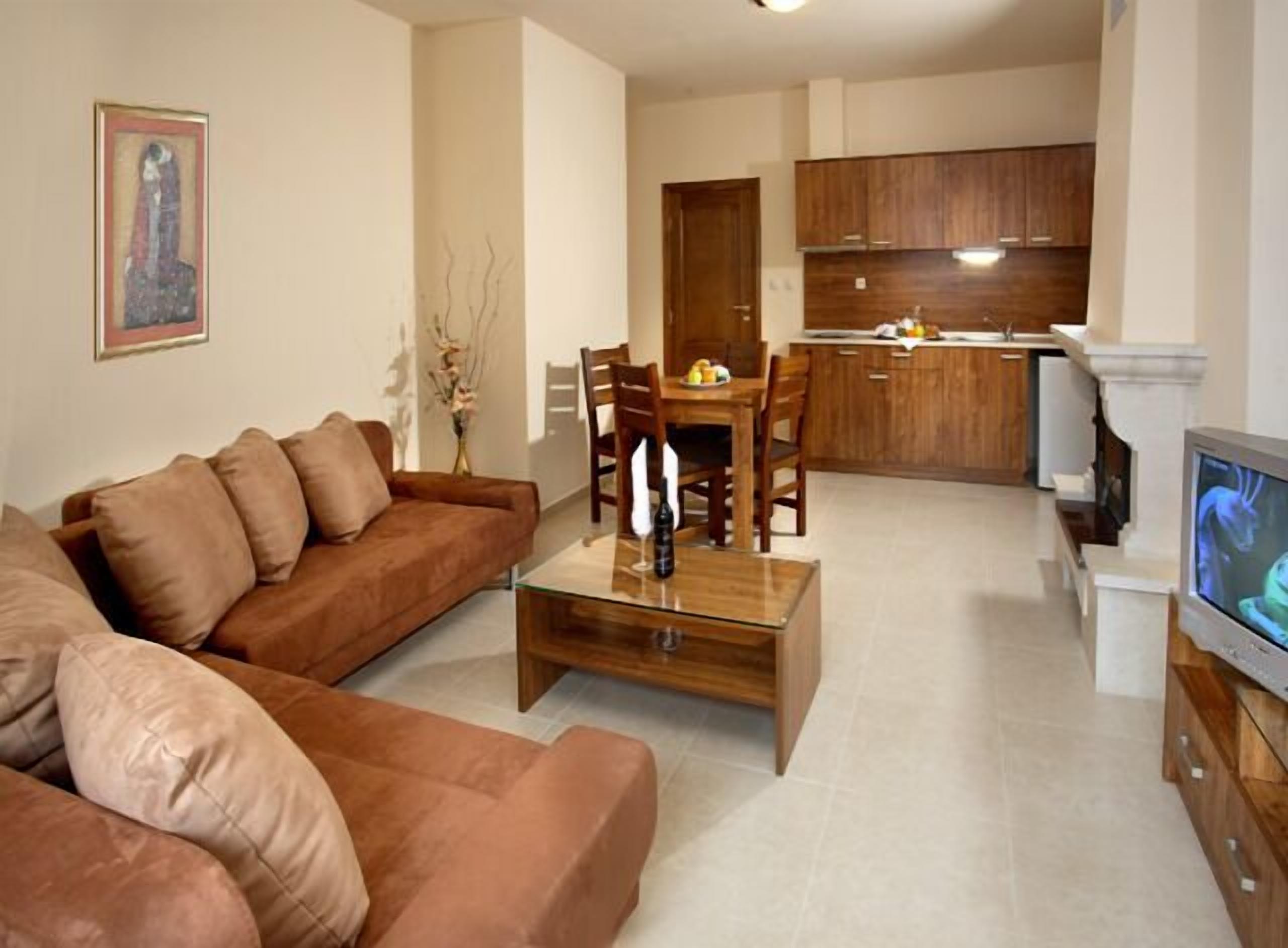 A living room with a brown sofa, a glass coffee table, and a dining area with wooden chairs and a small table. A kitchen with wooden cabinets is visible in the background.